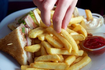Closeup fingers pick up a crispy fried potato wedge from a dish with pieces of toasted club sandwich ketchup and mayonnaise at the background