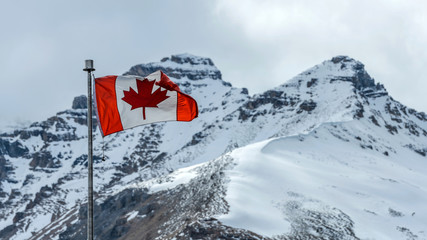 Canadian Rockies - A Canadian National Flag flying at front of two snow-covered high mountain peaks at Columbia Icefield Discovery Centre, Jasper National Park, Alberta, Canada.