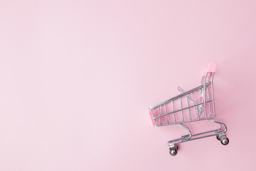 Small supermarket grocery push cart for shopping toy with wheels and pink plastic elements on pink pastel color paper flat lay background. Concept of shopping. Copy space for advertisement