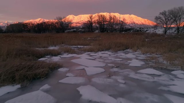 Aerial view of snow capped mountains at sunset in winter flying backwards over broken up ice on shoreline of Utah Lake.