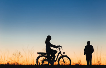 Obraz premium Silhouette of a girl and man on the e-bike or electric bicycle on the sunset background. Country style. Transportation in the village. Copy space. Female. Travel.