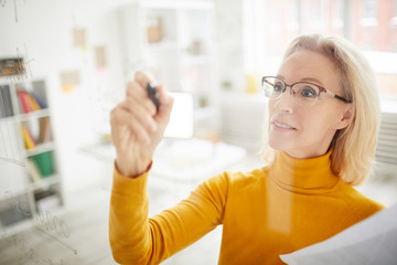 High angle portrait of mature businesswoman writing on glass wall in office while planning startup...