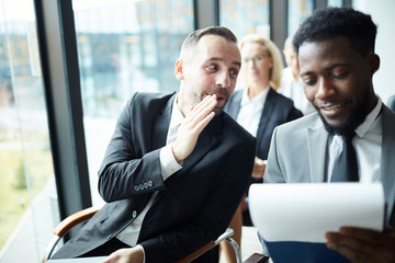 Young businessman whispering something to colleague sitting next to him while listening to report...