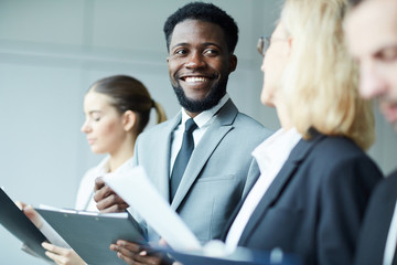 Young cheerful businessman with toothy smile looking at one of colleagues while discussing...