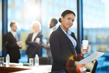 Young mixed-race businesswoman with paper and drink sitting in front of camera in working...