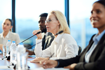 Confident blonde mature delegate in formalwear talking to microphone while looking at speaker