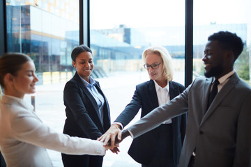 Four happy intercultural business people making pile of hands while standing in circle and looking...