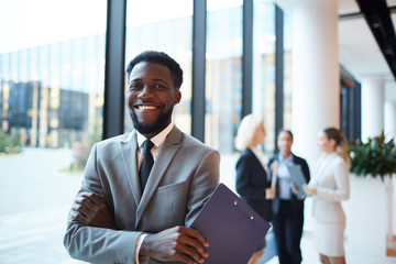 Young cheerful African-american entrepreneur or broker with toothy smile crossing his arms on chest in front of camera - obrazy, fototapety, plakaty