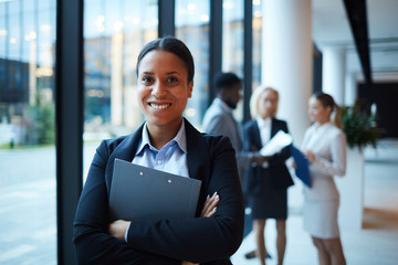 Young smiling mixed-race businesswoman with her arms crossed on chest looking at you on background of colleagues