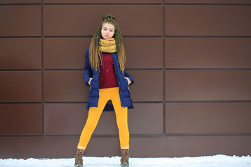 Portet of a young pretty white girl with a yellow knitted scarf in a blue jacket on the street...