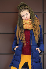 Portet of a young pretty white girl with a yellow knitted scarf in a blue jacket on the street opposite the modern brown background in winter.