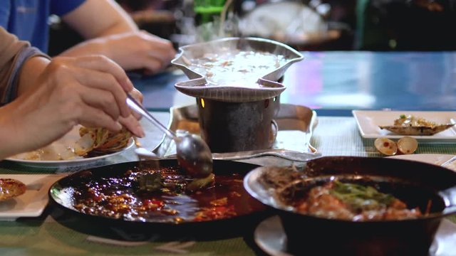 View across dining table, People eating various Asian foods, steaming bowls across scene, Hands taking different food choices.