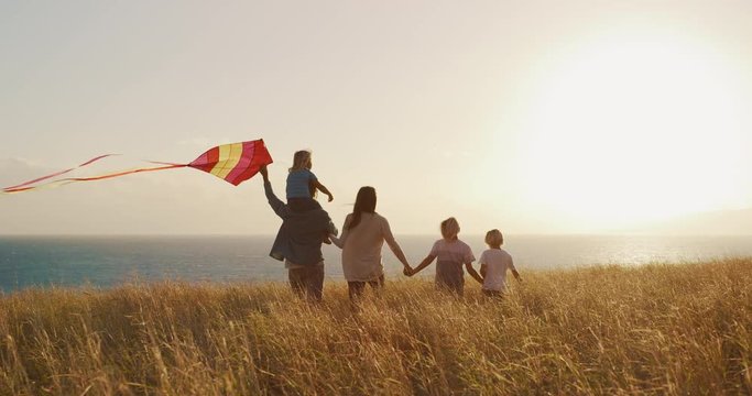 Happy family walking through fields of golden grass, flying kite, looking out to sea at sunset