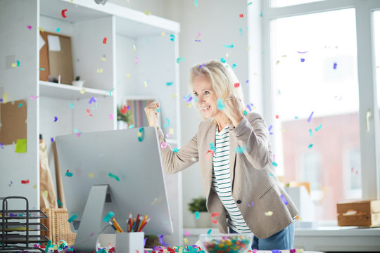 Portrait Of Excited Mature Woman Celebrating Success In Office And Looking At Computer Screen With Confetti Falling Overhead, Copy Space