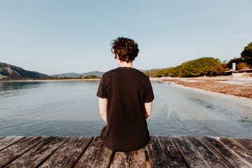 Junger Mensch von hinten sitzt im Sommer Urlaub auf einem Steg und genießt die Aussicht auf das Meer