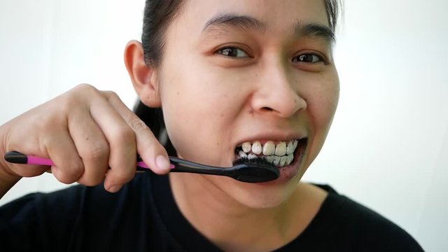 Asian young woman brushing her teeth with black toothpaste of activated charcoal, and black tooth brush on white background.