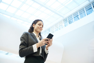 Young smiling businesswoman in formalwear reading message in smartphone in corridor of office center