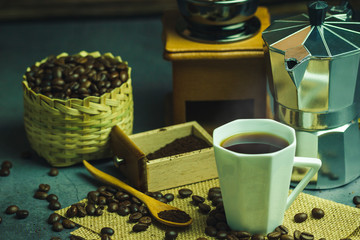 Brew black coffee in white cup and morning lighting. Roasted coffee beans in bamboo basket and wooden spoon. Vintage coffee grinder and pot. Concept of coffee time in morning or start the new day.