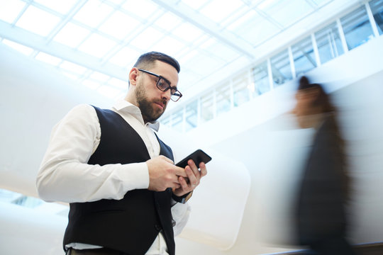 Young Serious Businessman With Smartphone Texting On The Move With Blurred Human Figure On Background