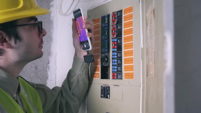 Electrician inspector using a thermal imaging camera attached to his smartphone as a tool to inspect the fuse box of the building.