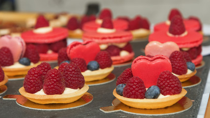 Raspberry tart cakes, tasty sweet desserts, baked dough baskets, cheese cream, top decorated by raspberries, blueberries and macron hearts. Street food market.