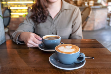 Closeup image of a hand holding a blue cup of hot black coffee with another latte coffee cup on wooden table in cafe