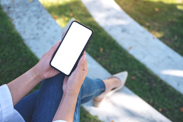 Mockup image of a woman holding black mobile phone with blank white screen while sitting in the outdoors