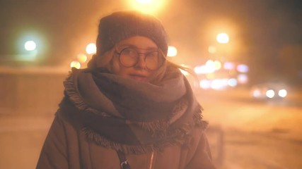Portrait of beautiful young blonde woman wearing fashion scarf standing in cold winter city street at night, looking at camera, closeup. Slow motion, shallow DOF, 4K UHD.