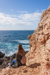 Happy female tourist in coat sits on cliff and enjoys sea view in Malta