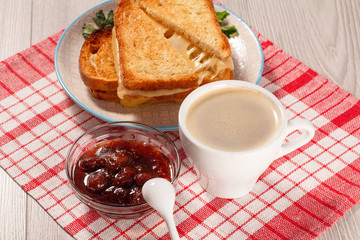Toasted slices of bread with cheese on white plate, cup of coffee, bowl with strawberry jam and spoon on napkin.