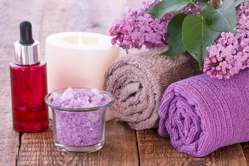 Lilac flowers, red bottle with aromatic oil, burning candle, bowl with sea salt and towels on wooden boards.