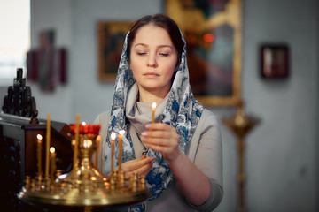 Russian beautiful Caucasian woman with red hair and a scarf on her head is in the Orthodox Church, lights a candle and prays in front of the icon.