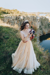 Fototapeta na wymiar Portrait of a beautiful bride in a beige dress with a crown on her head. A lovely bride with a bouquet is standing on a cliff in autumn weather. Wedding photography. Beautiful landscape.