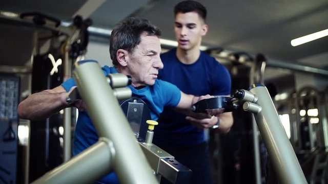 A senior man with a young trainer doing strength workout exercise in gym.