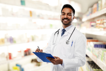 medicine, pharmacy and healthcare concept - smiling indian male doctor or pharmacist in white coat with stethoscope and clipboard over drugstore background