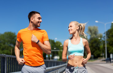 sport, technology and lifestyle concept - happy couple with fitness trackers running along bridge...