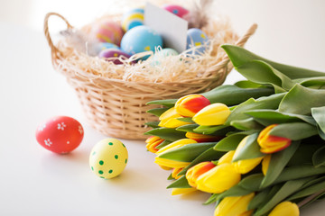 easter, holidays, tradition and object concept - close up of colored eggs in basket and tulip flowers on white background