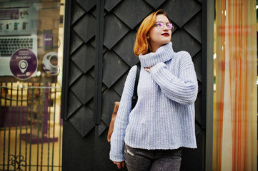Cheerful young beautiful redhaired woman in glasses, warm blue wool sweater with backpack posed outdoor.