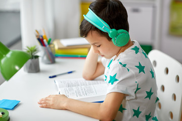 education, childhood and school concept - little student boy in headphones with textbook learning at home
