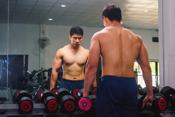 Fototapeta na wymiar An Asian man lifts a dumbbell in front of a mirror in a gym. Exercise in gym concept