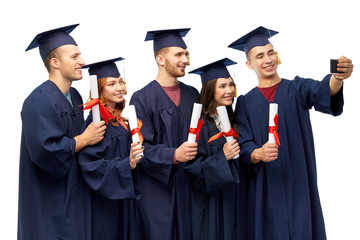 education, graduation and people concept - group of happy graduate students in mortar boards and bachelor gowns with diplomas taking selfie by smartphone over white background