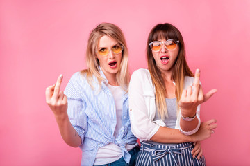 Fashion hipster girls. Pink background, not isolated.Two young woman showing middle finger,...