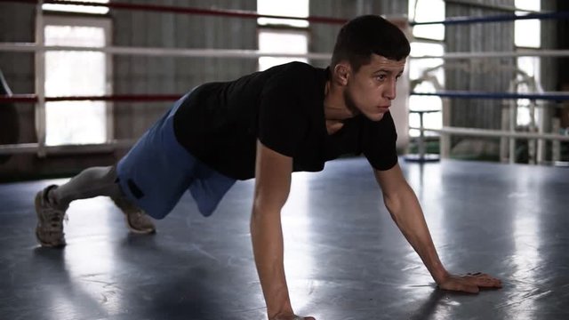 Close up of young muscular sportsman doing push-ups - from elbows to outstretched hands - while working out in boxing gym. Side view