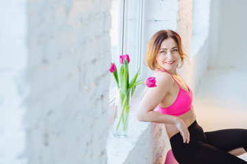 Mature elegant slim energy woman after good workout yoga pilates. Healthy lifestyle older age concept. Sports female in pink top black leggings relaxing near big window in white loft studio interior.