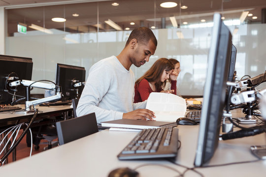 Serious male student reading book while sitting with friends at computer desk in library