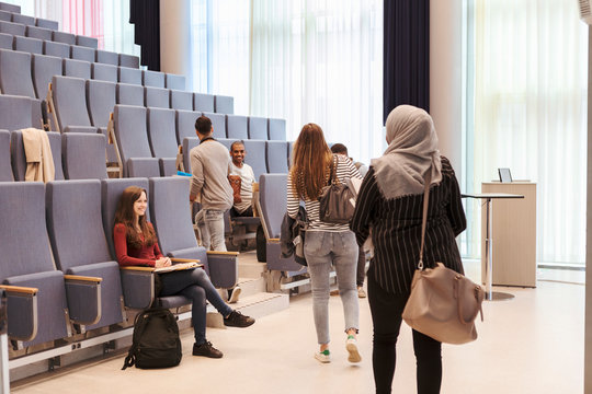 Smiling students looking at friends walking towards chairs in lecture hall at high school