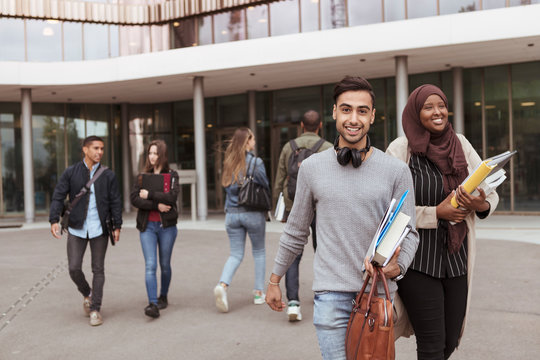 Portrait of smiling student holding books while walking in campus with friends in background