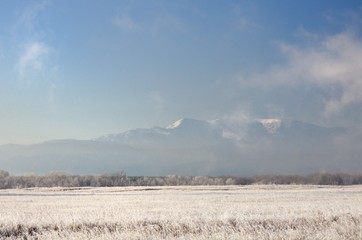 Winter landscape with snowy mountains behind field covered with frozen dry grass under dark blue...