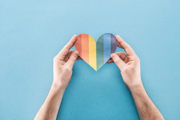 cropped view of male hands with rainbow colored paper heart on blue background, lgbt concept