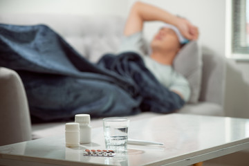 sick wasted man lying in sofa suffering cold and winter flu virus having medicine tablets in health care concept looking temperature on thermometer - obrazy, fototapety, plakaty